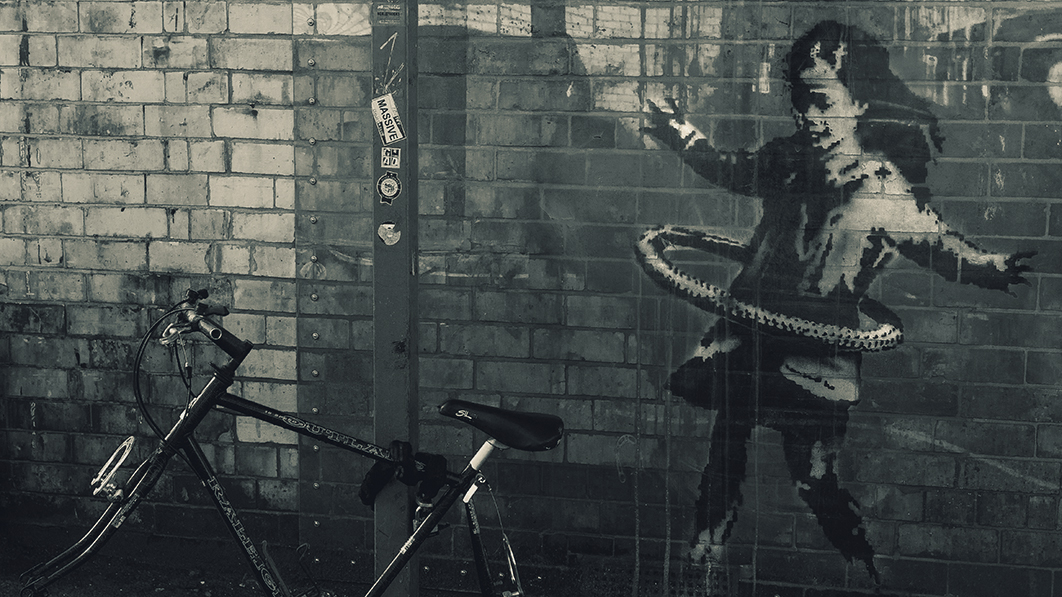 Foto einer grauen Ziegelwand mit angelehntem Fahrrad, auf der Wand: mutmaßlich ein Banksy-Bild: Mädchen mit Hula-Hoop-Reifen 